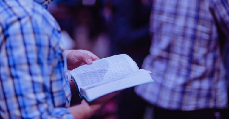 A man stands indoors reading a Bible, focusing amidst a crowd.