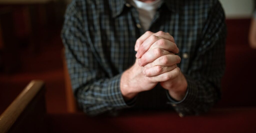 A man in a checked shirt praying in a church, hands clasped together.