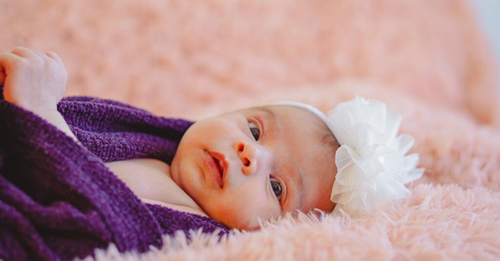 Adorable newborn baby with flower headband wrapped in soft purple blanket, lying on pastel pink bedding.