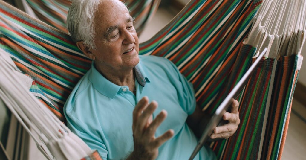 Elderly man enjoying leisure time in a hammock while using a tablet device.