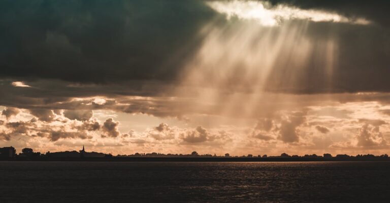 Sunbeams illuminate a serene landscape beneath dark storm clouds over a calm sea.