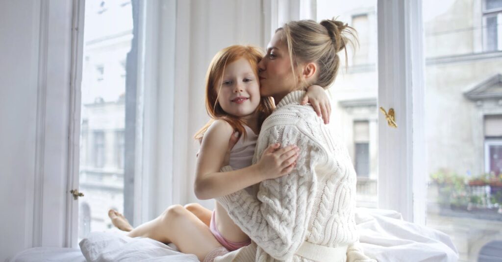 A mother affectionately embraces her daughter by a bright window, sharing a tender moment indoors.