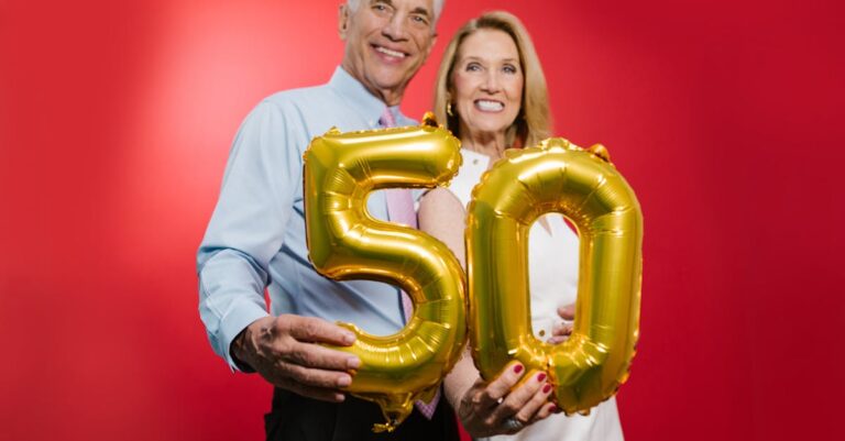 Happy senior couple celebrating 50th anniversary with golden balloons against a red studio backdrop.