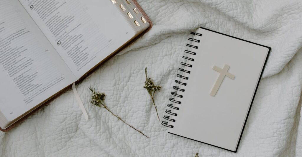 An open Bible and a cross-decorated notebook on white textured fabric with dried flowers.