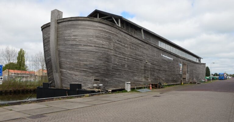 Large wooden structure by canal, resembling a historical ark on a cloudy day.