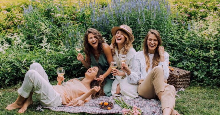 Four women enjoying a picnic, laughter, and wine in a vibrant summer garden.