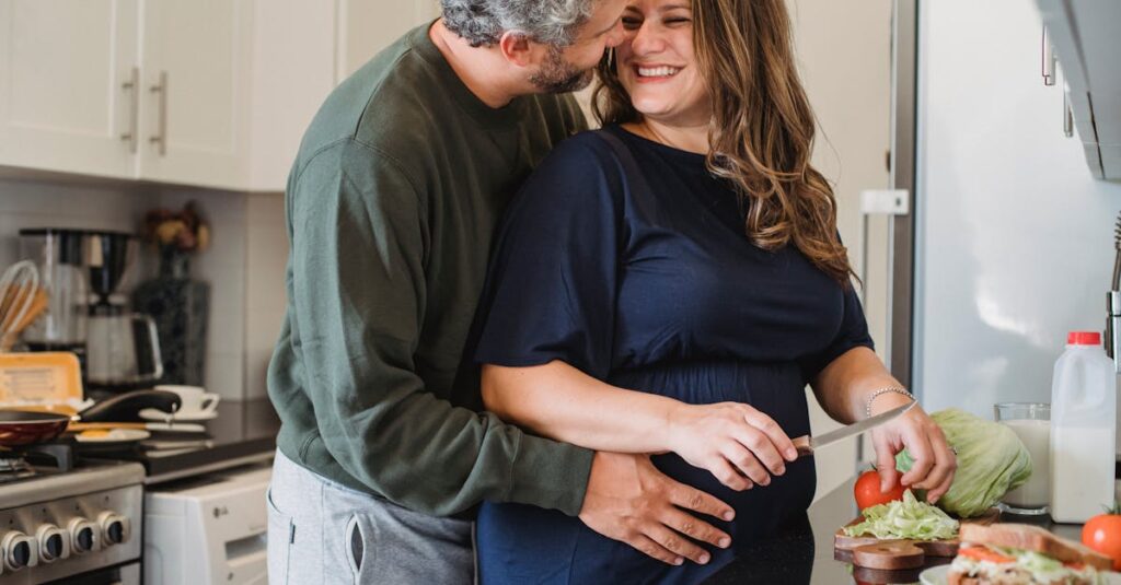 Cheerful pregnant wife in blue dress cooking sandwiches for breakfast while smiling and looking at husband embracing belly in modern kitchen