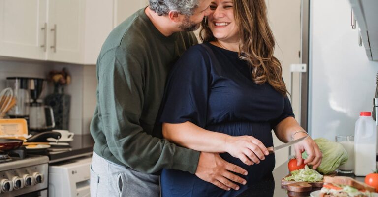 Cheerful pregnant wife in blue dress cooking sandwiches for breakfast while smiling and looking at husband embracing belly in modern kitchen