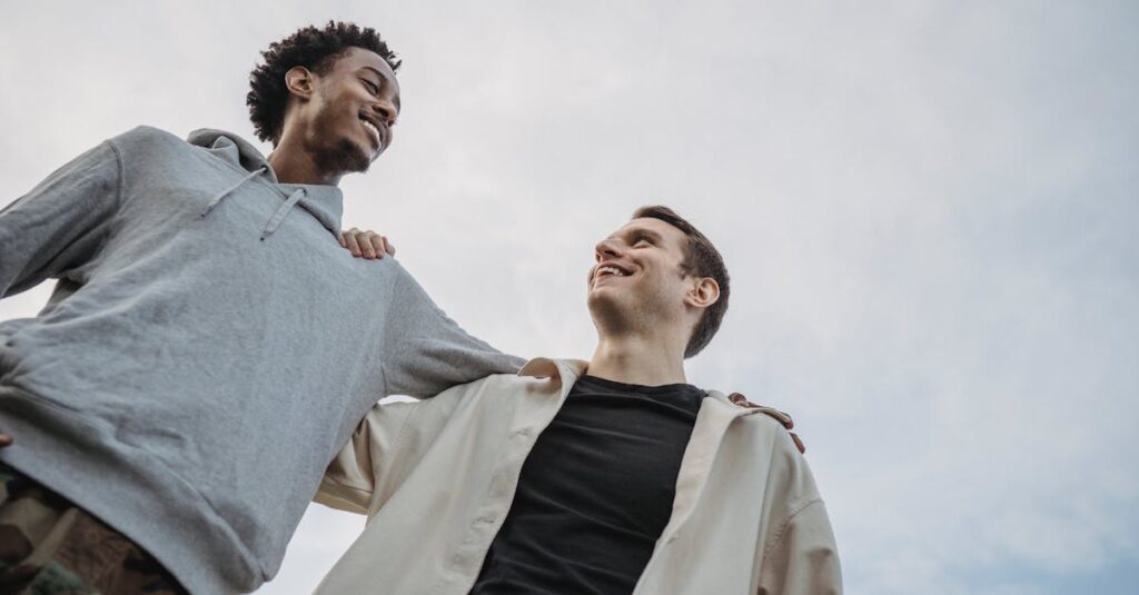 From below of cheerful young multiracial friends standing in hug looking at each other on cloudy day