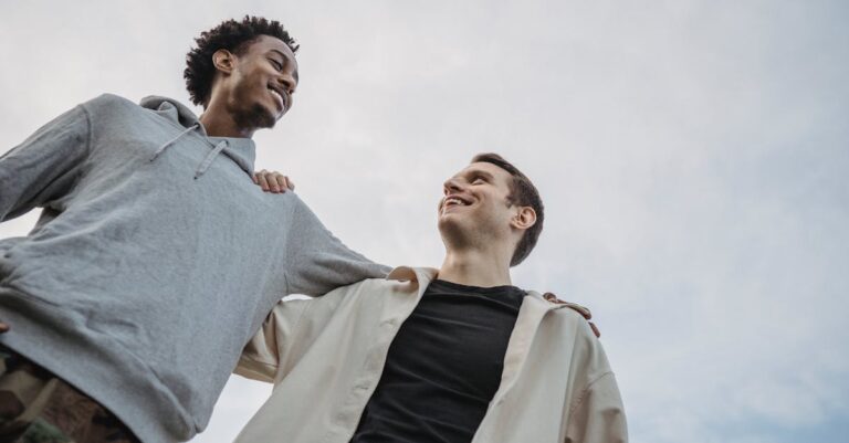 From below of cheerful young multiracial friends standing in hug looking at each other on cloudy day