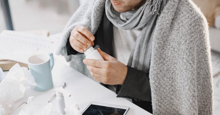 Adult male in scarf taking medicine at a desk with tissues and tablet, feeling unwell.