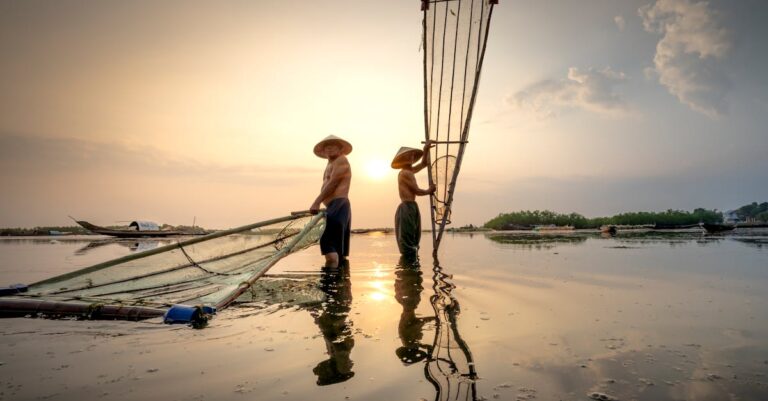 Asian fishermen in Vietnamese hats standing in river with fishing nets under cloudy sky in morning
