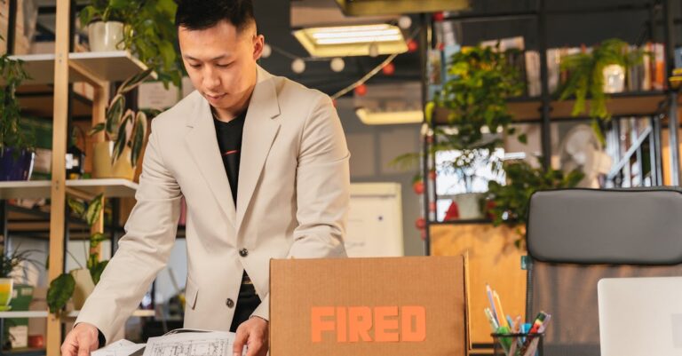An employee packing documents into a box labeled 'FIRED' in an office environment.