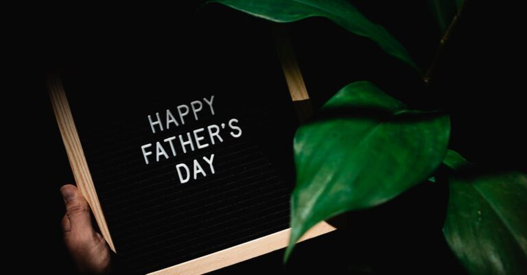 Dark background with Happy Father's Day sign held by a hand next to green leaves.