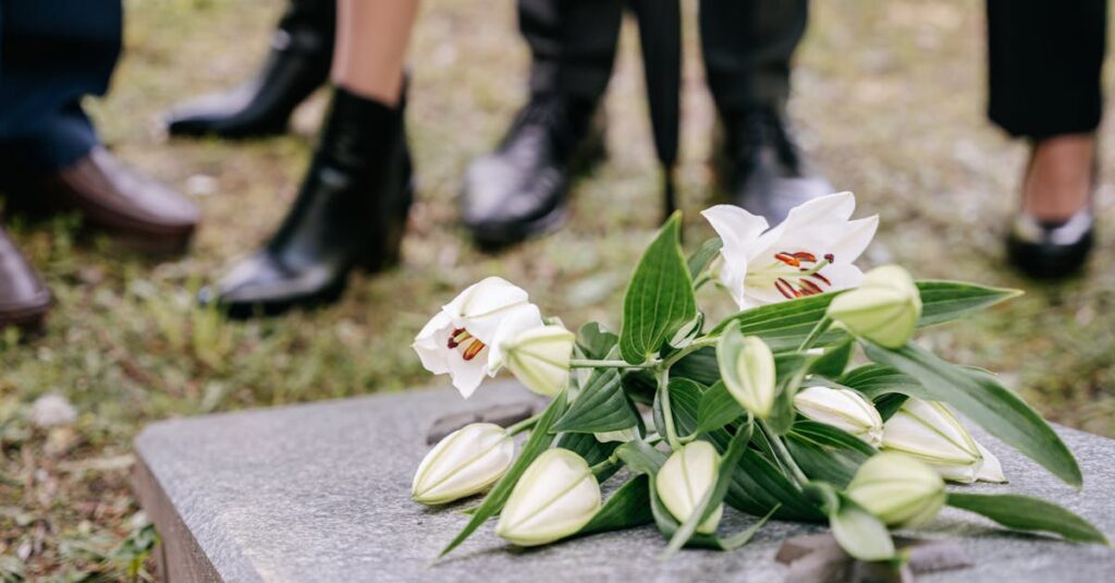 White lilies laid on a gravestone surrounded by people in mourning attire outdoors.
