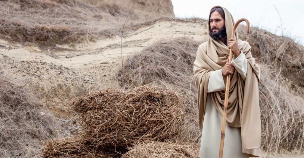 A person dressed in a biblical costume holding a staff beside hay bales in an arid landscape.