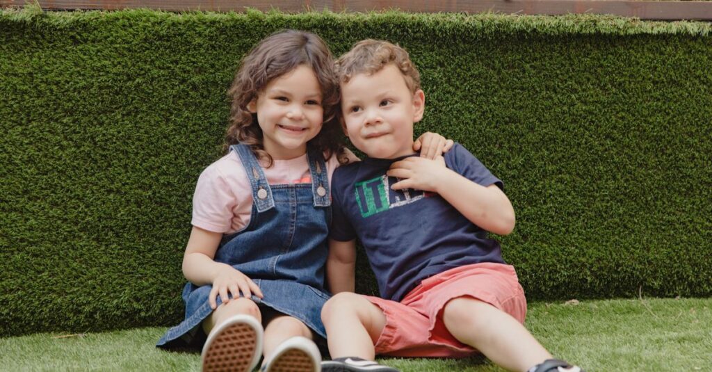 Two joyful children sitting and laughing on a vibrant green lawn, enjoying sunny outdoor fun.