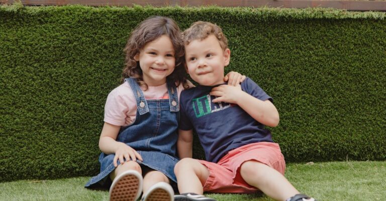 Two joyful children sitting and laughing on a vibrant green lawn, enjoying sunny outdoor fun.