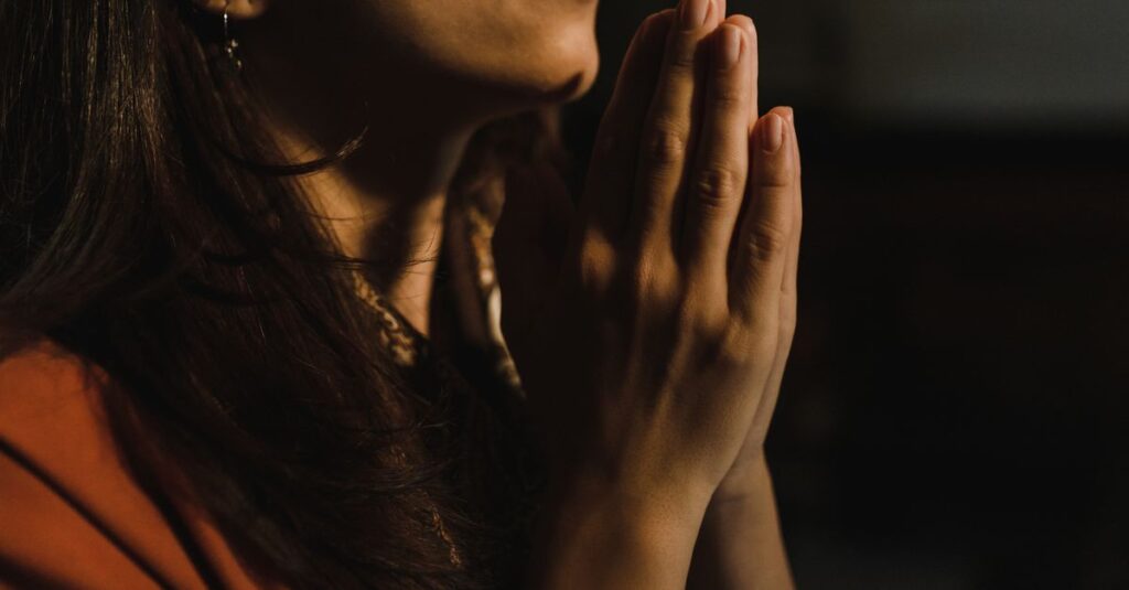 pexels-photo-5418299-5418299 Close-up of a woman with folded hands praying, captured in a serene indoor setting with low lighting.