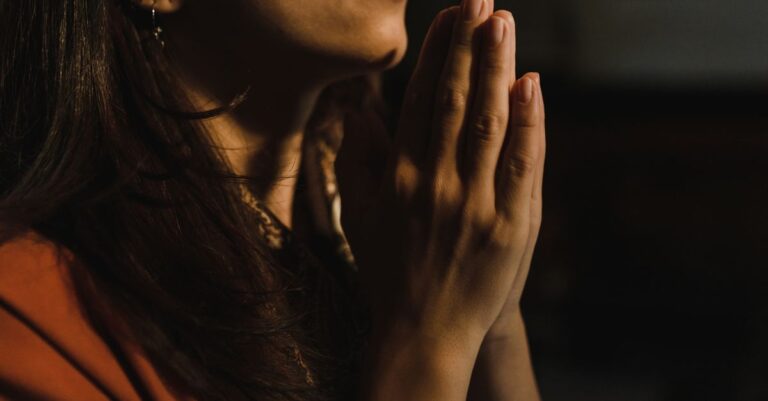 Close-up of a woman with folded hands praying, captured in a serene indoor setting with low lighting.