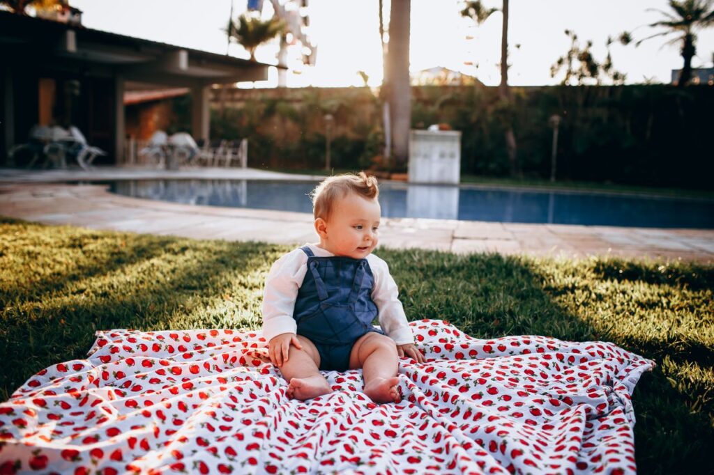 Adorable baby enjoying a sunny day on a strawberry-patterned blanket by a pool.
