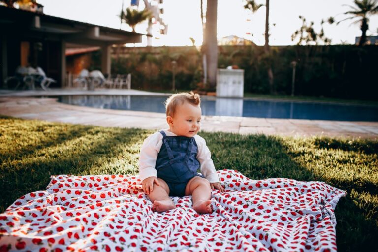 Adorable baby enjoying a sunny day on a strawberry-patterned blanket by a pool.