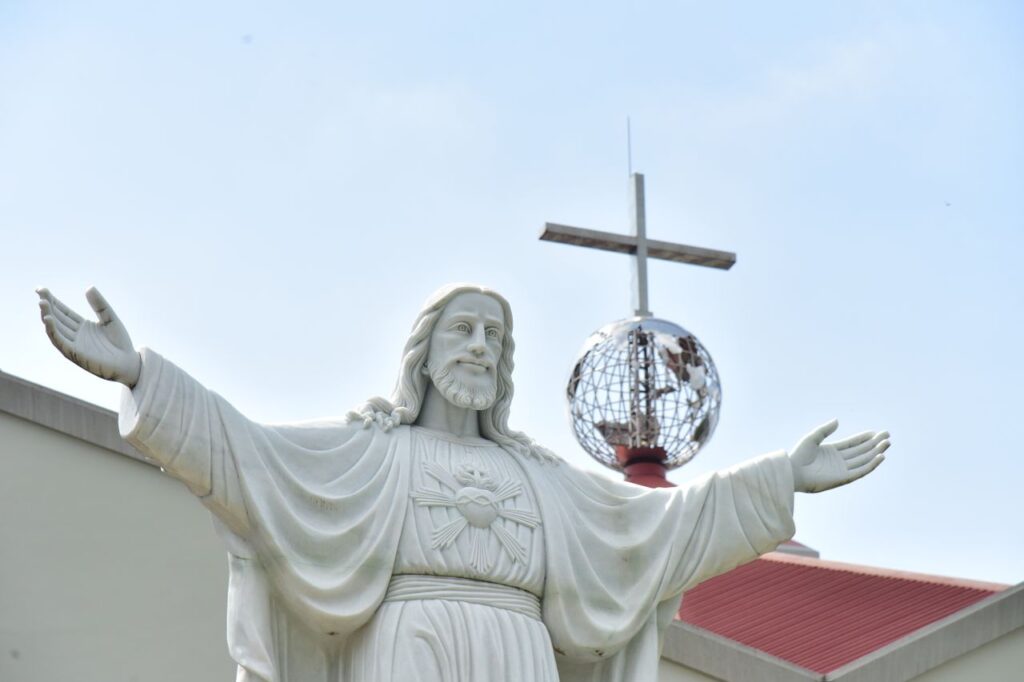 A white statue of Jesus Christ with outstretched arms in front of a building and cross.