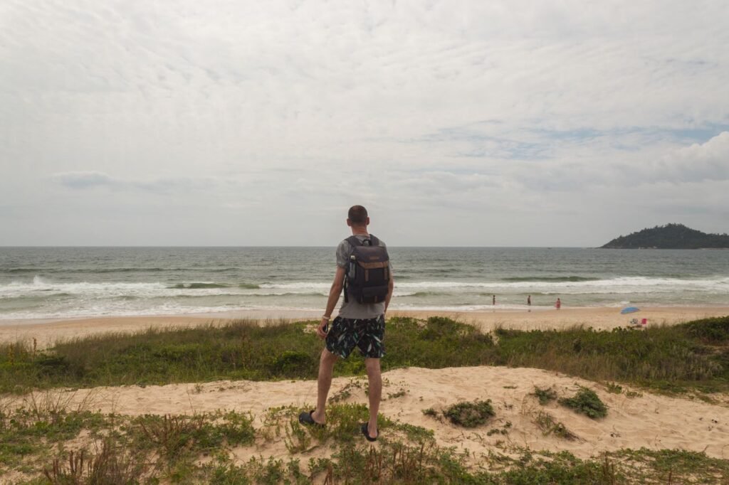 A man with a backpack stands on a hill overlooking a beach, capturing the serene ocean view.