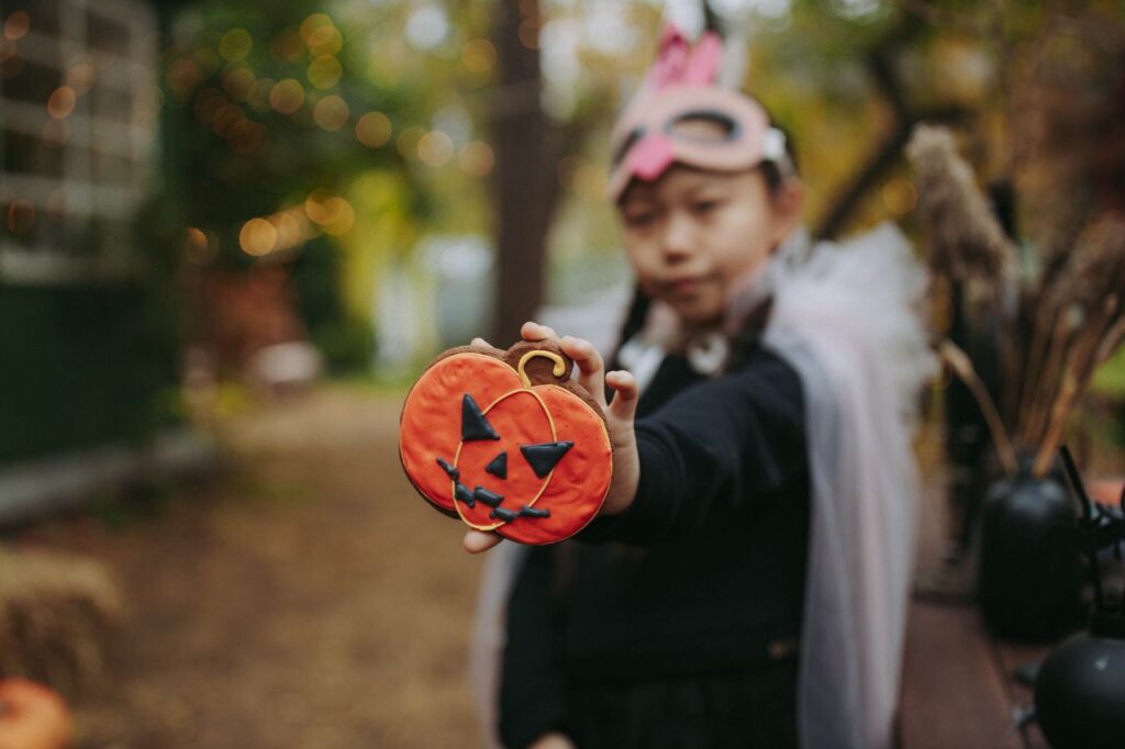 A child wearing a costume mask holds a Halloween-themed pumpkin cookie outdoors.