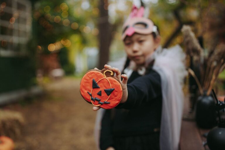 A child wearing a costume mask holds a Halloween-themed pumpkin cookie outdoors.