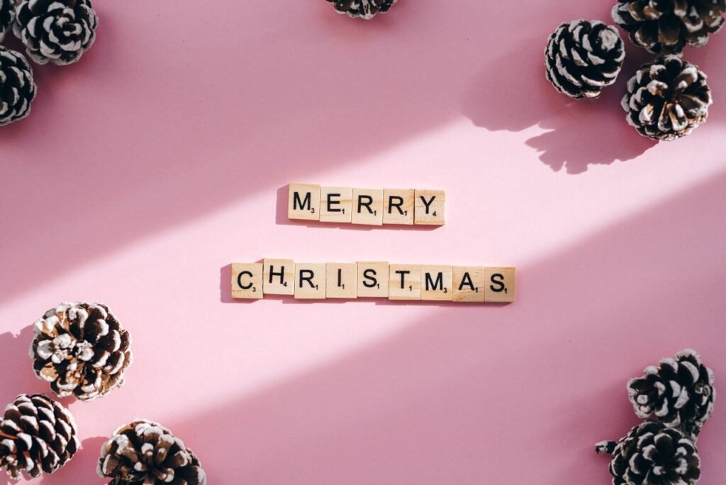 Top-down view of Merry Christmas spelled with Scrabble tiles surrounded by pine cones on a pink background.