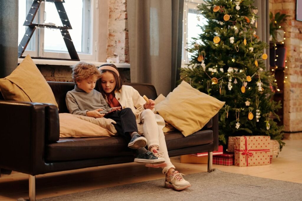 Two kids sitting on a sofa interacting with a tablet beside a decorated Christmas tree indoors.