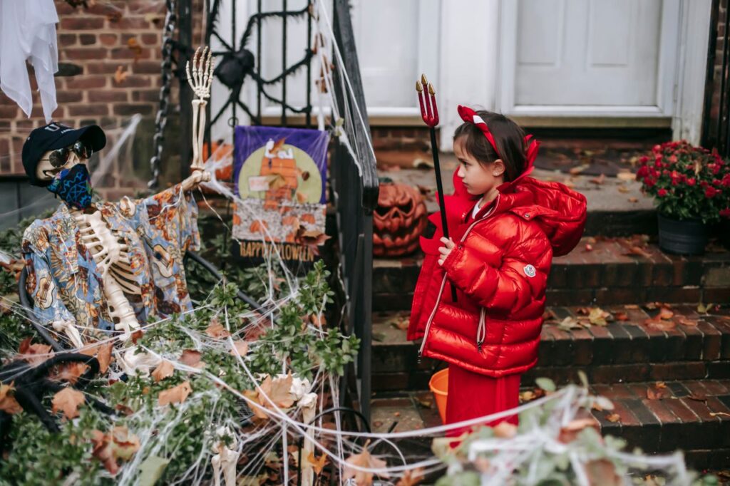 pexels-photo-5859401-5859401 Child in red devil costume with horns and pitchfork standing with bucket near skeleton and bushes in web on Halloween in autumn day outside