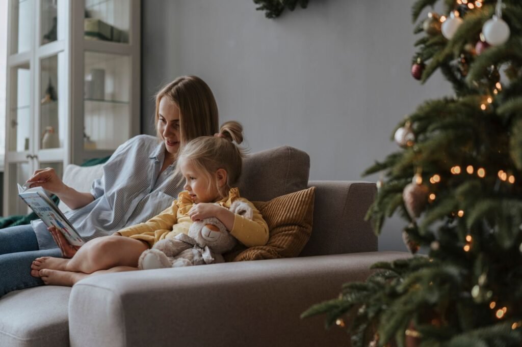 A mother and daughter sharing a book on the sofa next to a decorated Christmas tree indoors.