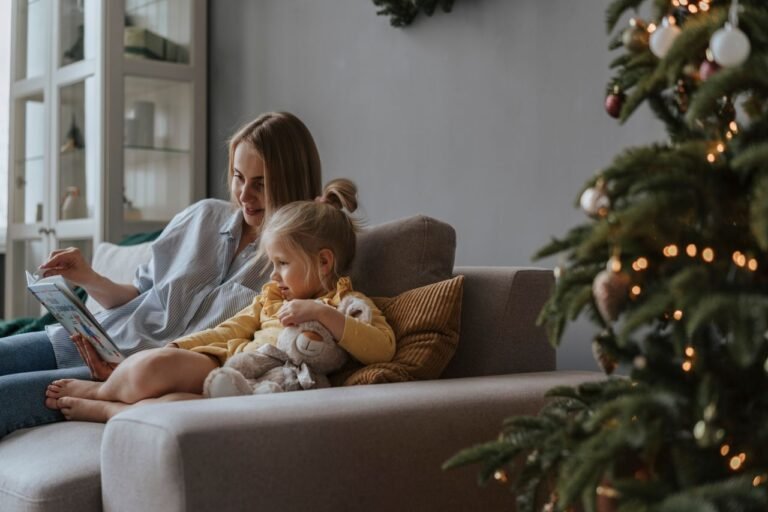 A mother and daughter sharing a book on the sofa next to a decorated Christmas tree indoors.