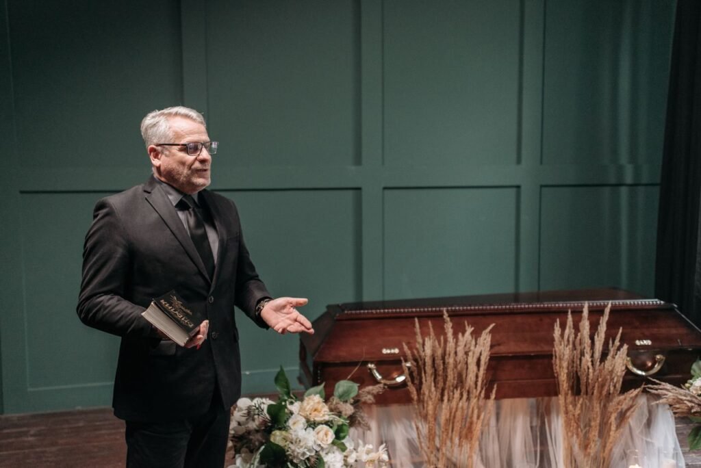 pexels-photo-7317717-7317717 A solemn funeral ceremony featuring a pastor in a black suit delivering a speech next to a wooden coffin adorned with flowers.