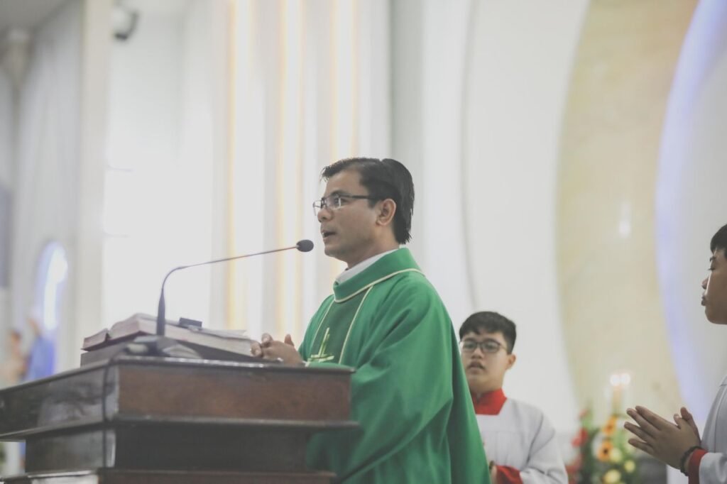 Priest in green robe giving a sermon during a church ceremony with altar servers nearby.
