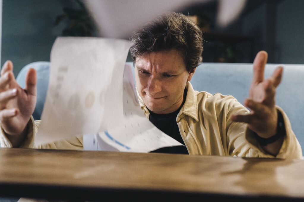 Stressed man throwing documents in frustration at a wooden desk in an indoor setting.