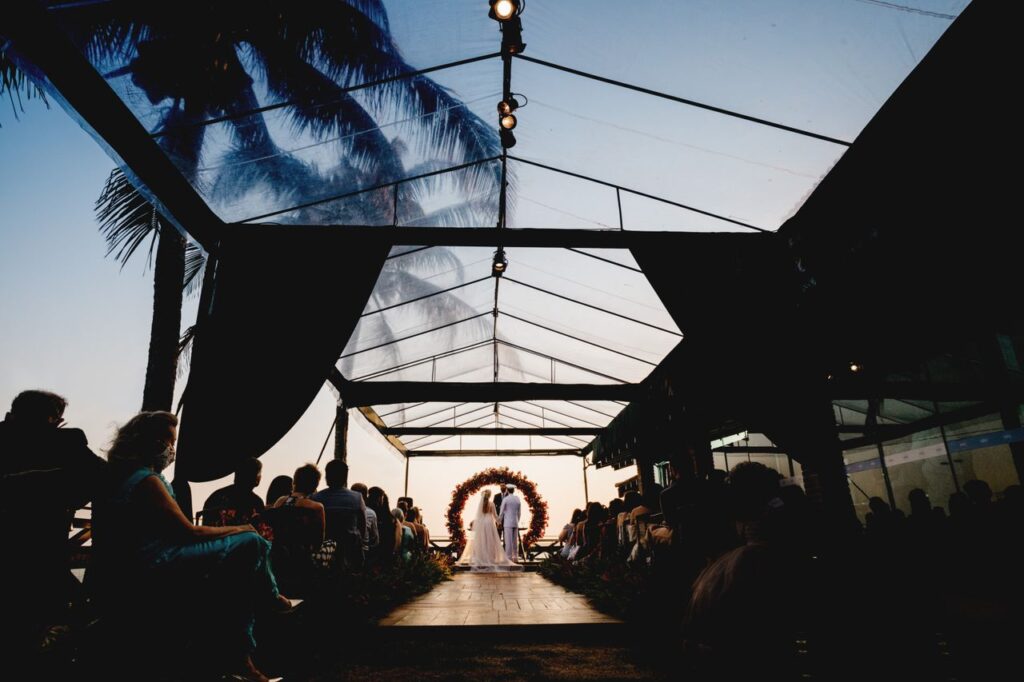 pexels-photo-9714792-9714792 Beautiful evening wedding ceremony under a glass tent with greenery and guests seated.